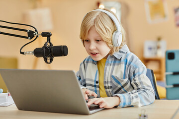 Cute blond boy in headphones looking at laptop screen and speaking in microphone during livestream...