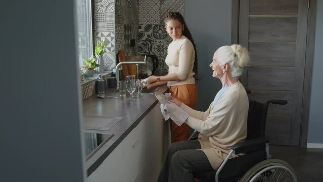 Assisted living nurse doing dishes while elderly woman in wheelchair drying plates with towel in kitchen at home