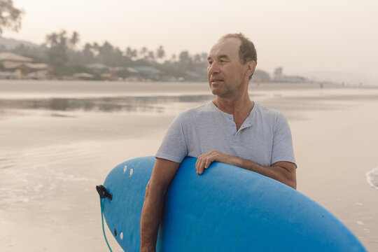 Confident mature surfer walking along empty shore at sunset, carrying blue surfboard under armpit and looking aside.