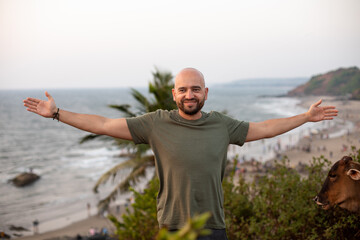 Happy man standing with arms wide open on green hill in front of seashore against seascape and...
