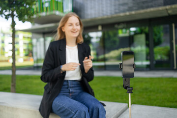 Female blogger recording video outdoors. Beautiful young girl smiling and looking at the camera standing in the summer park, talking and gesticulating with both hands. Blogging, video blog and people
