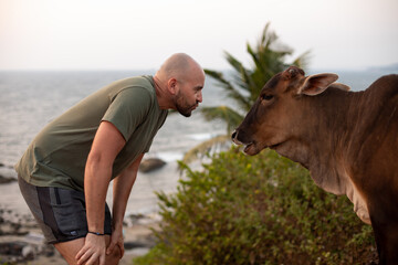 Friendly vacationist leaning towards humped cattle standing against seascape and meeting brown...