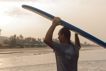Strong middle aged man carrying blue long surfing board on head coming out of sea and looking at...