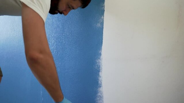 caucasian bearded man painting wall with paint roller. Painting apartment, renovating home with blue color paint. High quality FullHD footage