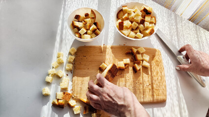 Homemade cooking and cutting of slices of diced dry white rye bread on cutting board and white plate. Hand of woman with Crackers for food, healthy eating on table in sunny day, top view