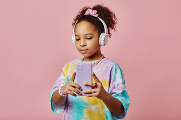 Cute and pretty African American child in headphones taking selfie while looking at smartphone camera against pink background in isolation