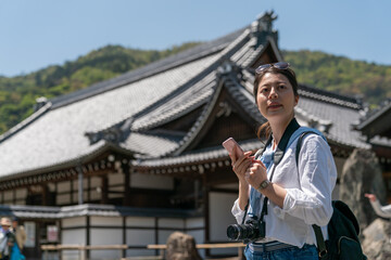 asian Japanese female visitor looking into distance at historic Buddhist buildings at Tenryuji temple while checking travel info on phone in Arashiyama Kyoto japan