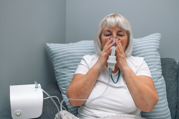Sick upset caucasian senior woman with an inhaler. Unhealthy female doing inhalation at home, she...