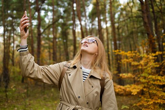 Woman in a style beige coat stands in a autumn forest on a hike and catches a phone call on a smartphone with his arm raised up. Hiker looking for a mobile network on a smartphone. High quality photo