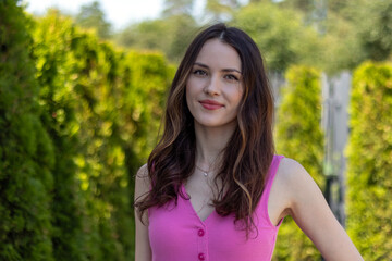 Fototapeta premium Portrait of a beautiful young woman model with wavy brunette hair in a pink dress against a background of green bushes
