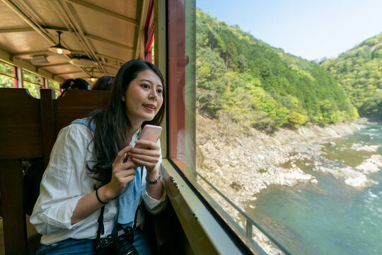 cheerful asian Korean girl tourist enjoying natural scenery of Arashiyama Mountains out of window as she tries to take photos with cellphone during the sagano romantic train ride in Kyoto japan