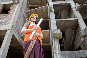 Portrait of confident young beautiful Indian female civil engineer or architect wearing helmet and...