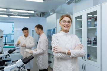 Fototapeta premium Portrait of a young woman scientist, chemist, doctor standing in the laboratory of the clinic, crossing her arms on her chest and smiling at the camera. In the background, male colleagues.