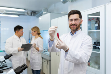 Portrait of a young male doctor standing in the laboratory with a flask of blood in his hands, pointing at the camera and smiling. In the background, colleagues are discussing research.