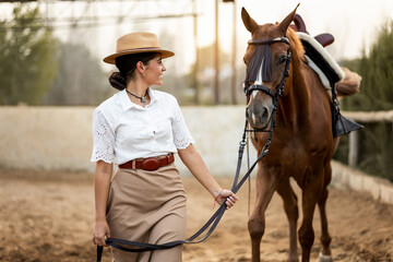 Half-length photo of a pretty girl in fancy clothes walking her brown horse. The woman pulls the reins of the Algo-Arabian animal while looking at it. Concept of women riding horses.