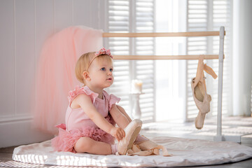 A cute little ballerina in a pink ballet costume sits near the barre in the room and tries to put on her pointe shoes. Kid and ballet dance. Copyspace.