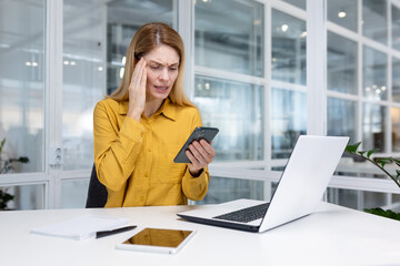 Mature experienced business woman using app on phone online sitting inside office at workplace with laptop, female worker shocked by bad news reading online message from smartphone.