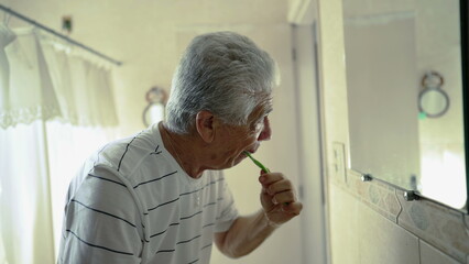 Elderly man brushes teeth in front of bathroom mirror reflection, morning daily ritual, depicting...