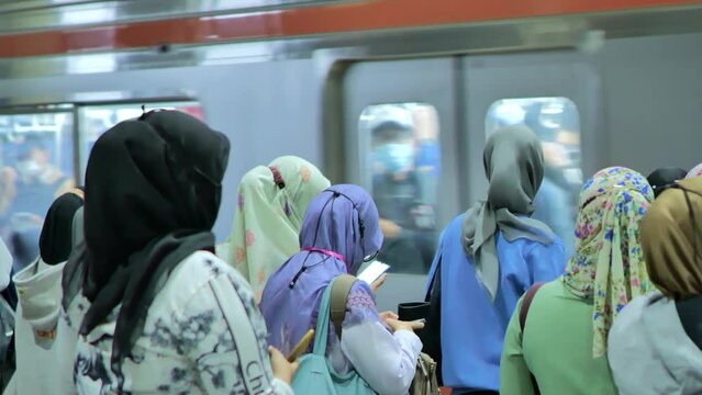 The female passengers are standing and using their phones beside the commuter train coming in at station in Jakarta city at night. Depicts the boredom of train passengers at the station after work.