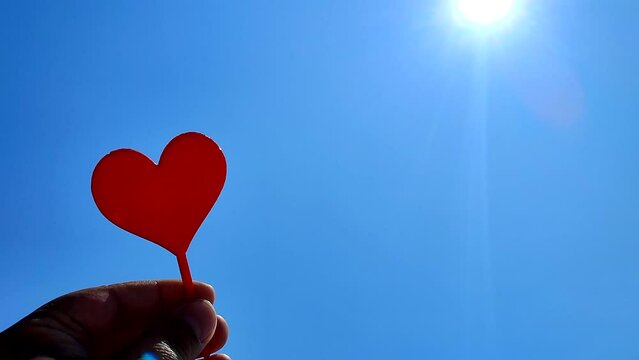 Person holding stick in shape red heart in hand on background of blue sky with shining sun on sunny summer day. Concept love relationship romance amour celebration St Valentines Day