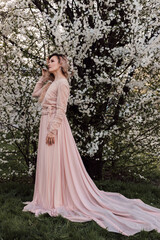 A woman in long pink dress stands near cherry blossom. Spring Blooming Garden.