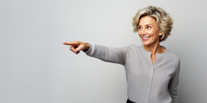 a happy, smiling, mature classy fashionable Caucasian woman, pointing finger to the side - on light gray background with copy space