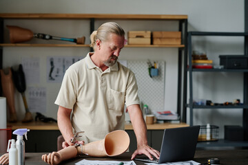 Side view portrait of senior man making arm prosthetics in workshop and using computer, copy space