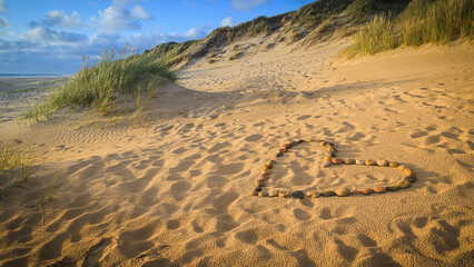 Ein Herz aus Steinen liegt an einem Sandstrand mit Dünen am Meer