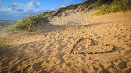 Zelfklevend Fotobehang Ein Herz aus Steinen liegt an einem Sandstrand mit Dünen am Meer © mpix-foto