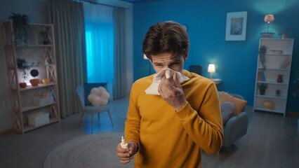 A man with symptoms of a cold stands in the living room close up. A man blows his nose into a paper handkerchief and holds a nasal spray. Home medicine concept. - obrazy, fototapety, plakaty