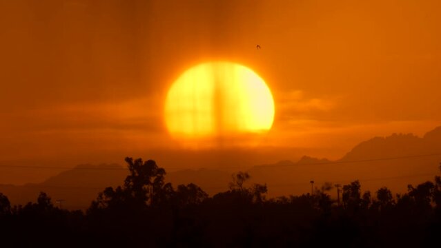 This super telephoto video captures a glowing sun setting over mountains with a rain storm in the foreground.