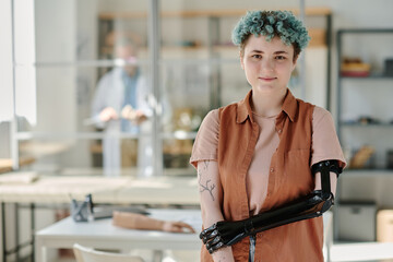 Waist up portrait of young woman with bionic prosthetic arm posing in doctors office and smiling at...