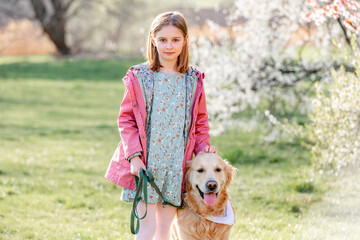 Girl with golden retriever dog walking outdoors in spring park with blossom trees. Pretty female child kid with pet doggy labrador at nature in sunny day