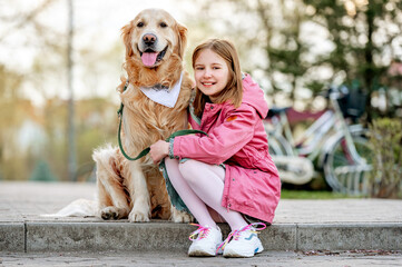 Girl with golden retriever dog outdoors in sunny day portrait. Female child kid hugging pet doggy labrador at street
