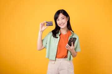 Experience the excitement of mobile shopping with a vibrant young Asian woman 30s, donning orange shirt and green jumper, using smartphone to show credit card on yellow studio background.