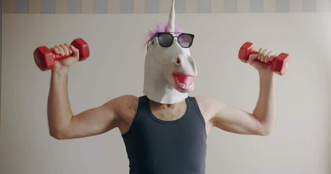 Funny man with unicorn mask and sunglasses doing exercise with weights for hand training on the white background at home. Comical fitness at home. Humorous stretches. Funny home workout.