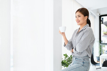 Young business woman standing to drinking coffee and thinking about the strategy of new startup while looking out the window in modern office. copy space