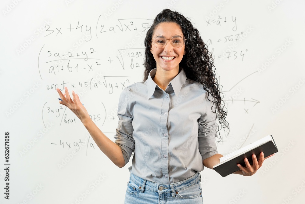 Wall mural Portrait of a beautiful Oriental curly-haired female student, teacher, tutor standing in front of blackboard with math tasks. Smiling girl greets the audience holding a notebook in her hands.