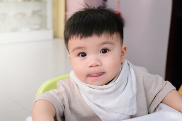 Close-up portrait of asian baby sticking out tongue with messy mouth sitting indoors on the baby feeding chair, baby eating concept