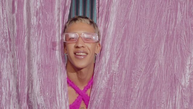 Trendy queer guy with makeup wearing feminine outfit peeking through pink curtains on stage, looking from side to side and smiling on camera