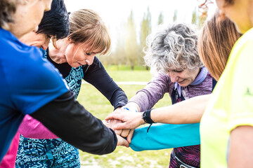 Happy multigenerational women stacking hands together before sport workout outdoor - Female friends having fun together outdoor at city park - Bright filter with focus on grey hair woman