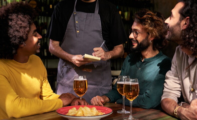 Multiethnic group of three male friends in a bar ordering some food to an unrecognizable waiter who is taking notes. Copy space.