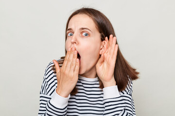 Shocked woman with brown hair wearing striped casual shirt standing isolated over gray background keeps hand near ear covering mouth with palm listening gossips.