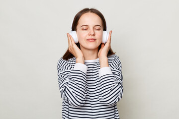 Satisfied woman with brown hair wearing striped casual shirt standing isolated over gray background enjoying favorite tracks with closed eyes keeping hand on headphones.
