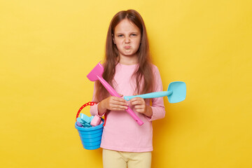 Sad unhappy bored little cute girl with dark hair holding beach sandbox toys rake and shovel isolated over yellow background looking at camera with pout lips grimacing expressing negative emotions.