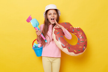 Sunny coast adventure. Excited amazed little cute girl in panama holding beach sandbox toys rake shovel and rubber ring isolated over yellow background screaming making announcement. - obrazy, fototapety, plakaty