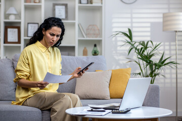 Dissatisfied and disappointed woman behind paper work sitting on sofa at home, Hispanic woman...
