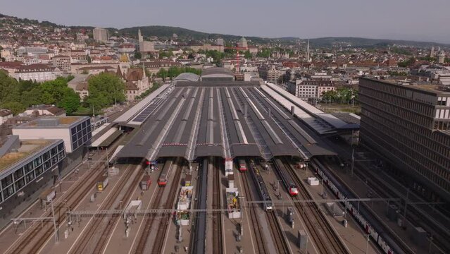 Forwards fly above main train station. Roofed platform and passenger trains on tracks. Aerial view of railway infrastructure in city. Zurich, Switzerland