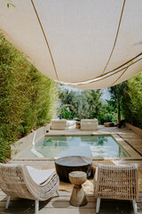 A tranquil outdoor lounge with a private pool, surrounded by lush greenery and shaded by a canvas canopy. Cozy wicker chairs and neutral tones create a relaxing, nature-inspired retreat