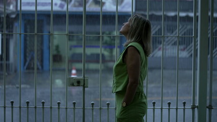 Pensive Woman Observing Rainy Afternoon in Neighborhood from Garage, Looking Through Metal Bars at...